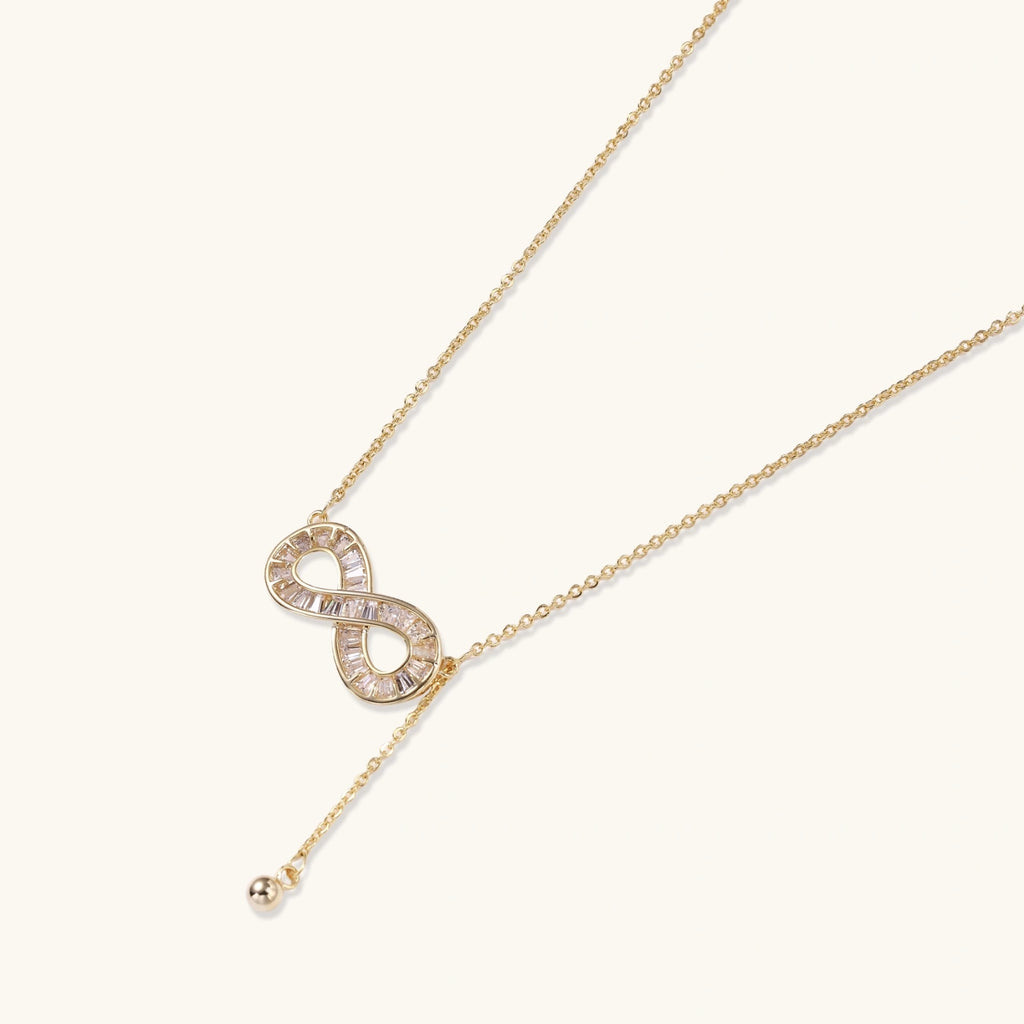 Gold infinity pendant with baguette crystals on adjustable chain against cream background.