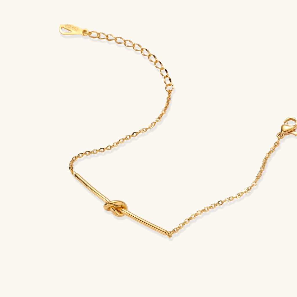 Delicate gold chain bracelet with minimalist bar and centered knot detail on cream background