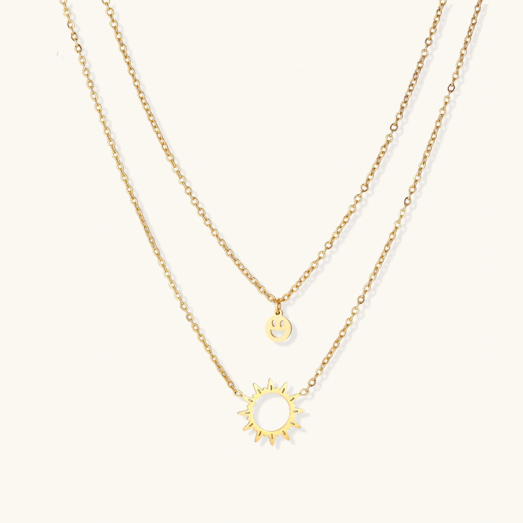 Gold layered necklace set with sunburst and smiley face pendants on delicate chains against white background.
